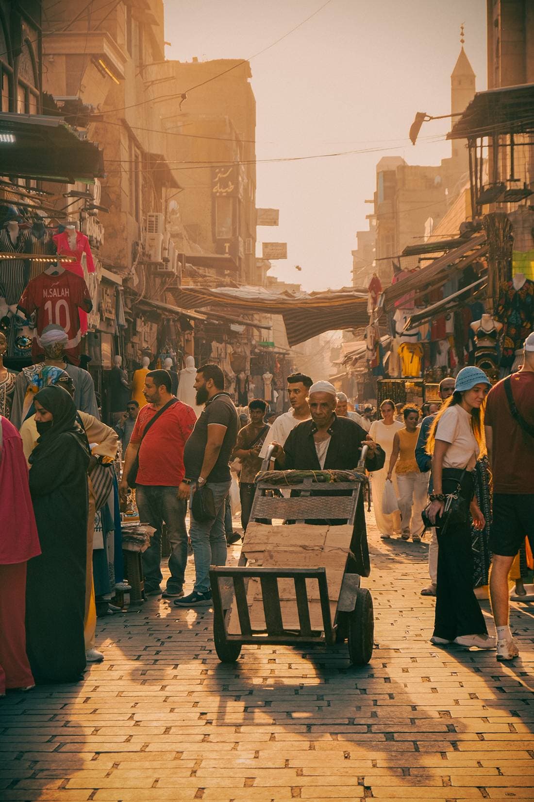 A busy market street in warm evening light, with a handcart in the foreground