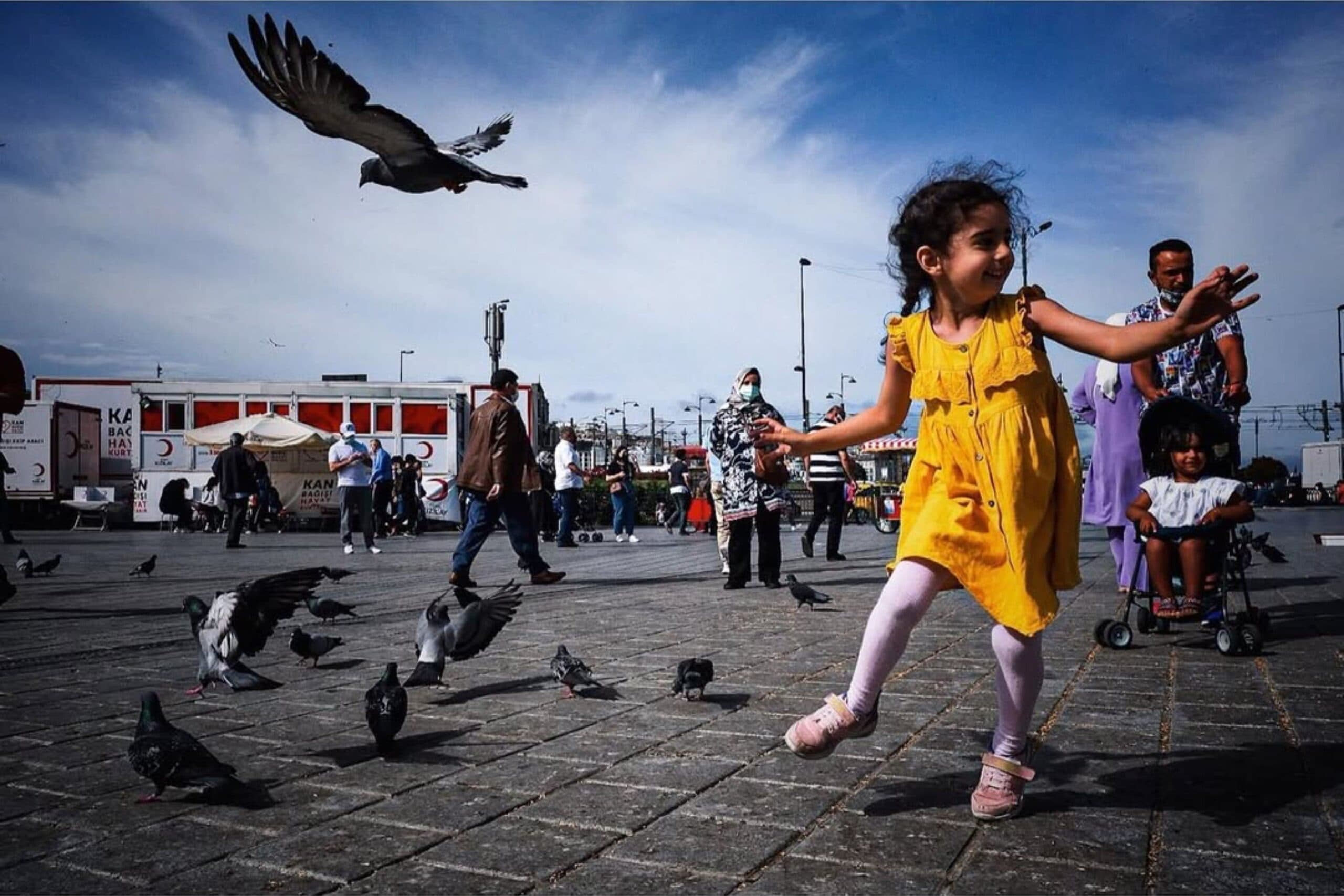 A child in a yellow dress moving among pigeons in a busy public square