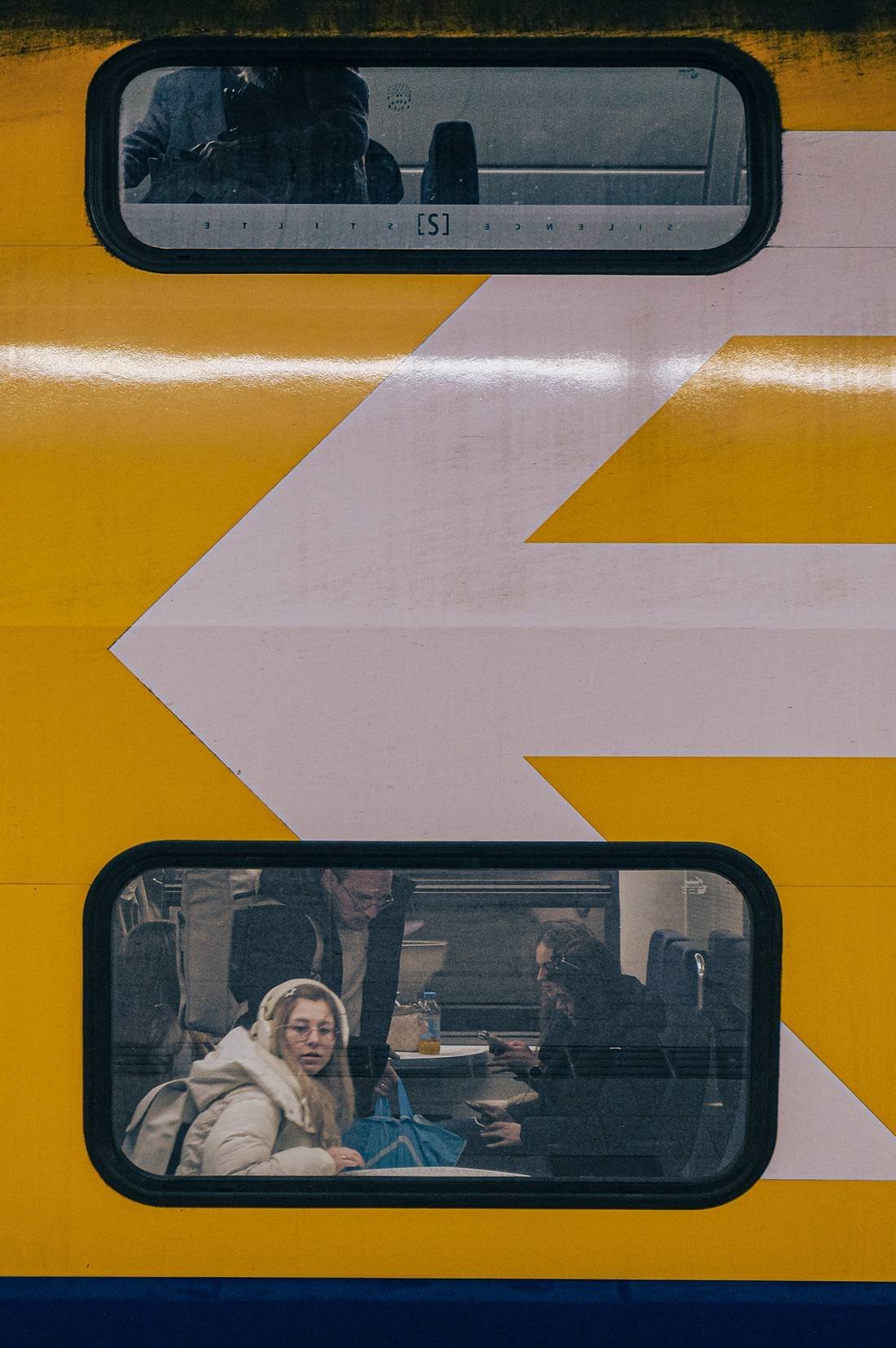Passengers seen through two windows of a yellow train divided by a large painted arrow