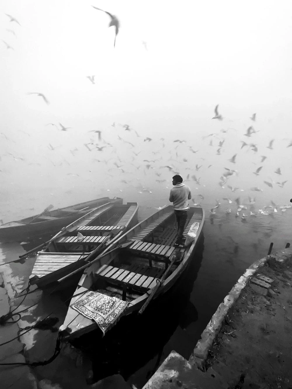 Empty boats and a lone figure surrounded by birds in fog, photographed by Priya Ojha