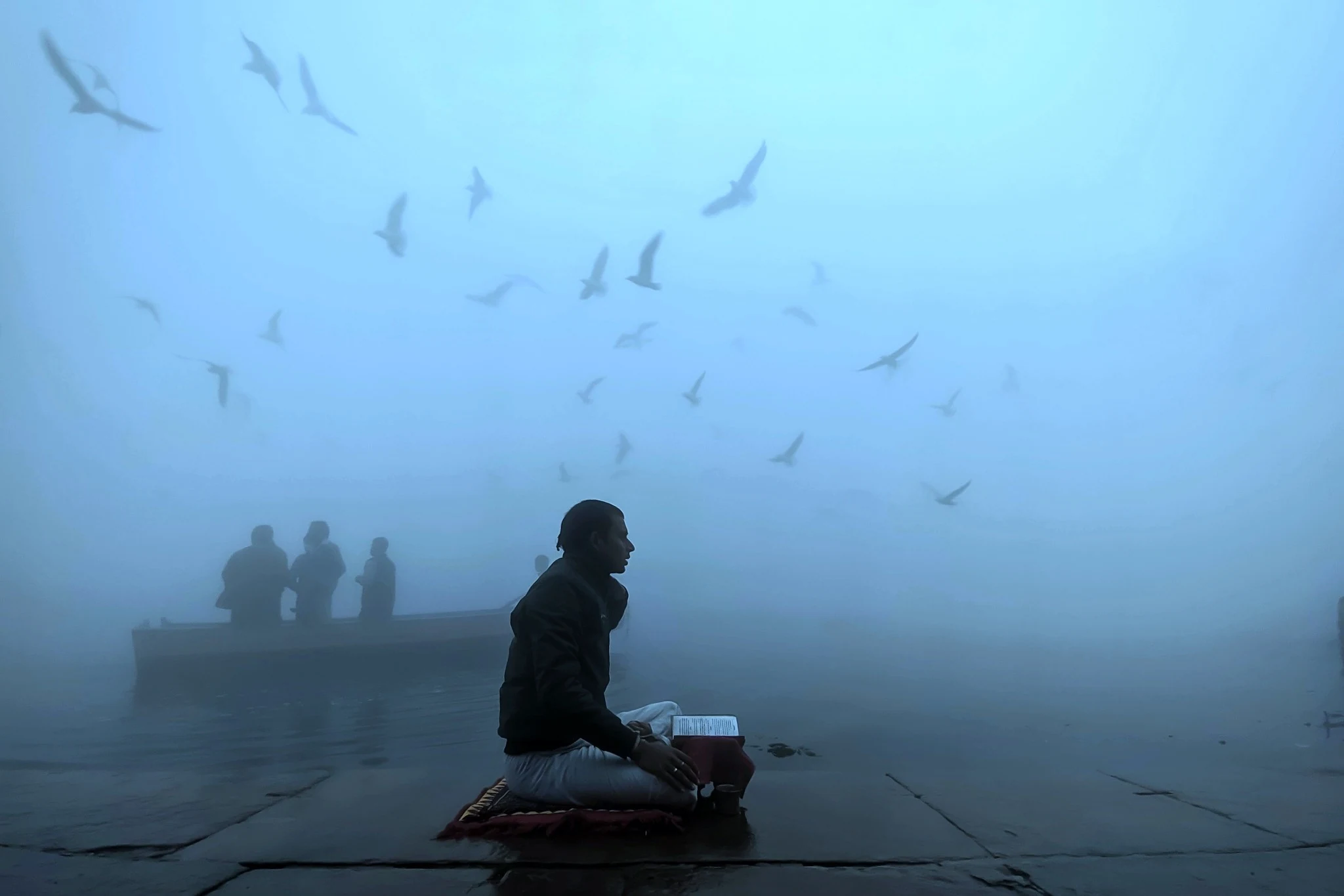 A seated man in morning fog beneath a flight of birds, photographed by Priya Ojha